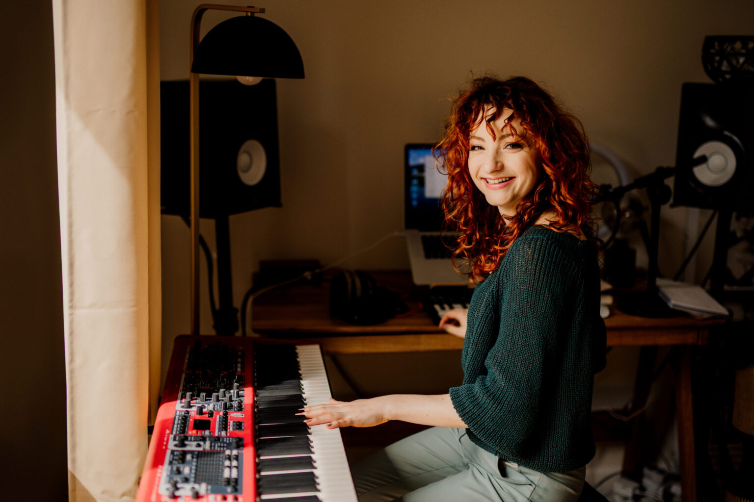 Redheaded singer and vocal coach smiling to the camera while being in her voice studio; with a red piano and monitors.